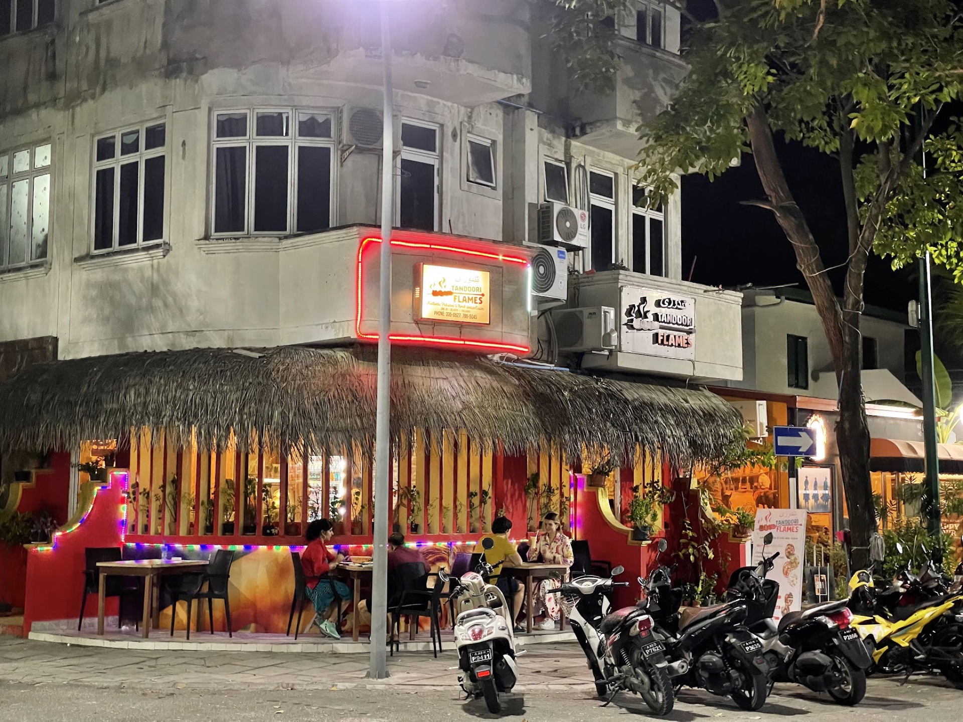 Tandoori Flames corner at night, neon sign lit above the thatched terrace